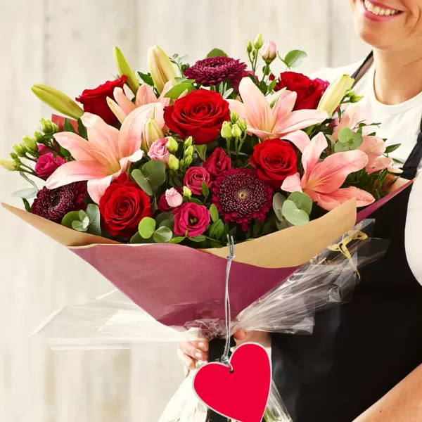 Buchet de flori cu trandafiri roșii și crini roz, ținut de o persoană, cu etichetă roșie în formă de inimă