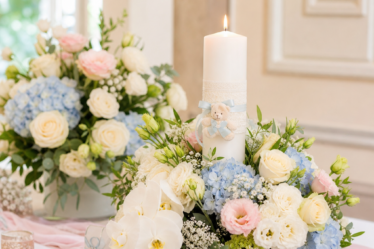 aranjamente florale botez - Fotografie luminoasă cu aranjamente florale de botez în nuanțe pastel, pe o masă elegantă, cu flori albe, roz pal și bleu, verdeață fină și o lumânare d